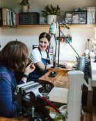 A lifestyle scene in a jewelry workshop showing a crafter smiling while working at a wooden craft table with a jeweler's loupe and tools, with another person seated in the foreground.