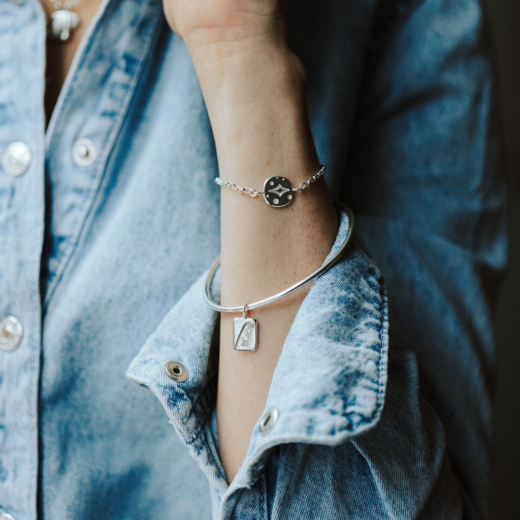 Woman in a denim shirt wearing a bangle featuring a feather charm and a bracelet featuring a star charm on her wrist.