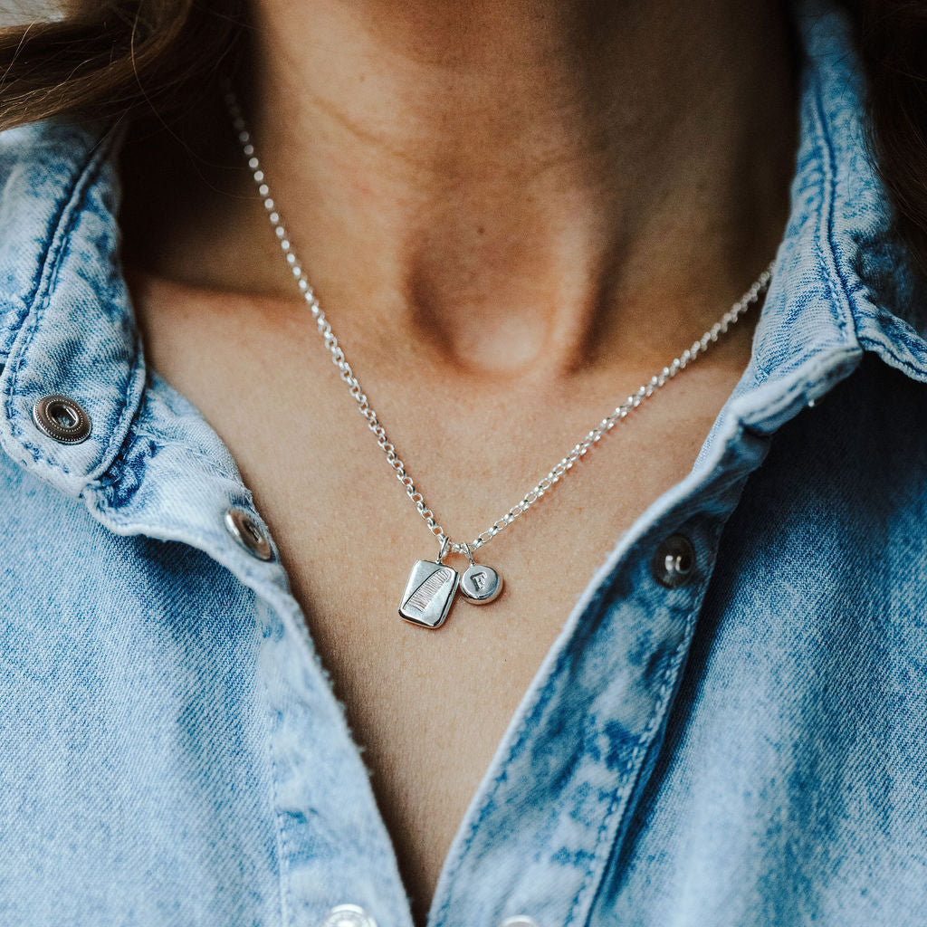 Woman in a denim shirt wearing a charm necklace, one charm with a feather design stamped on and the other has the letter F.