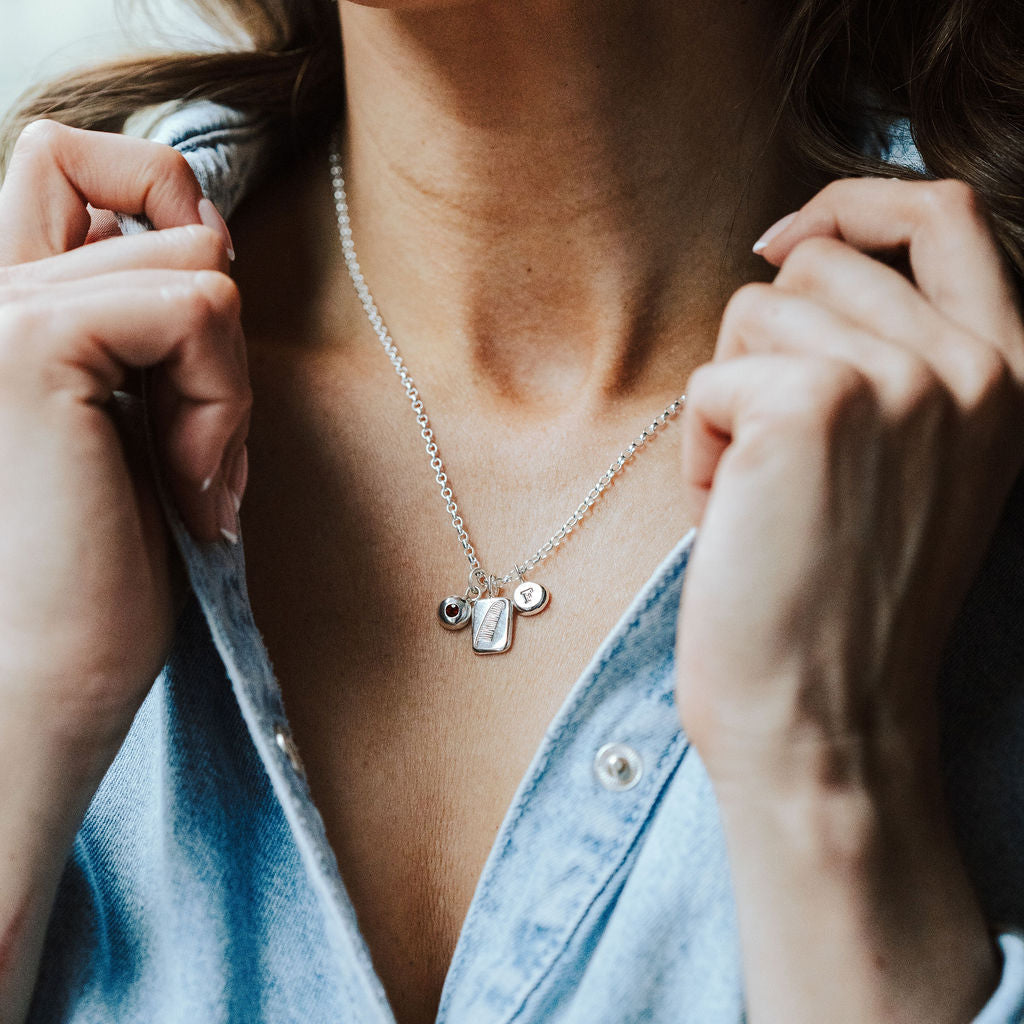 Woman wearing a denim shirt and a charm necklace featuring a feather charm, initial charm and birthstone charm.