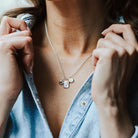 Woman wearing a denim shirt and a charm necklace featuring a feather charm, initial charm and birthstone charm.