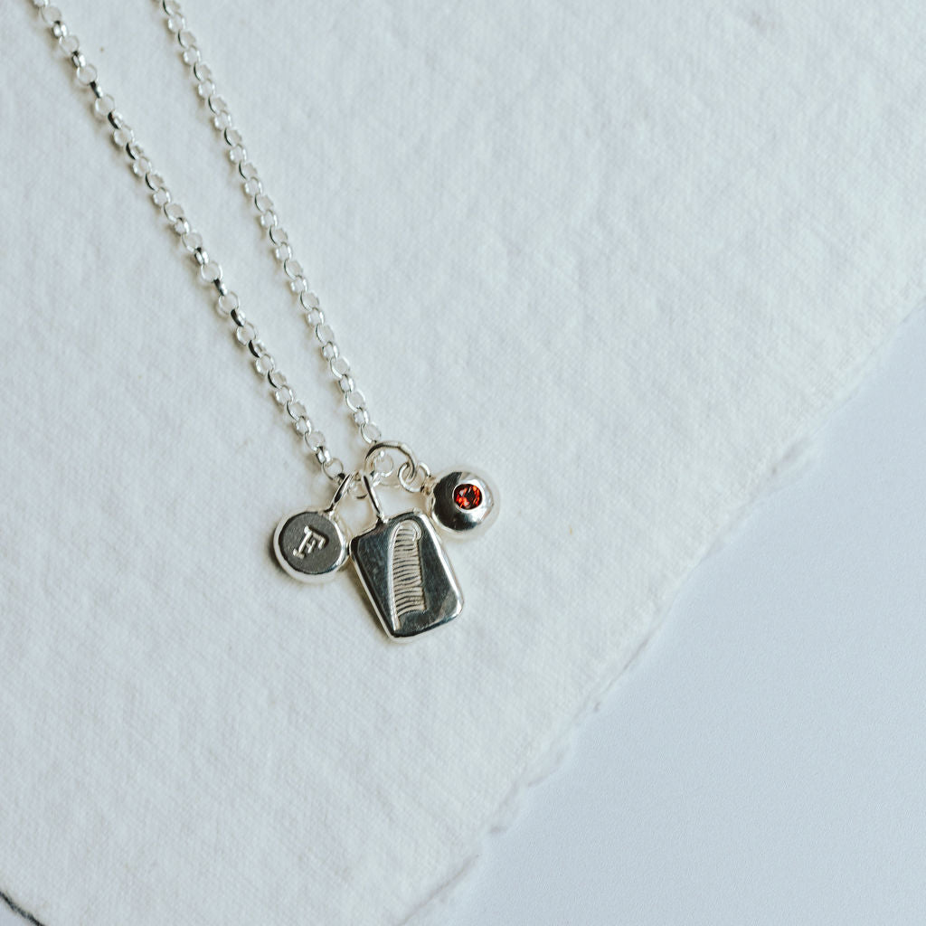 Necklace with three charms on a white background, featuring a feather design, and initial and a birthstone charm.