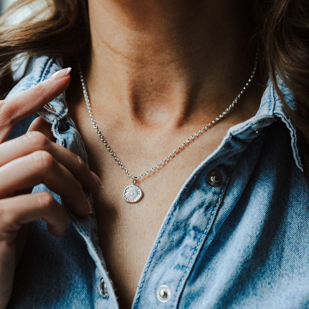 Woman in a denim shirt wearing a necklace with a sunrise design on it.
