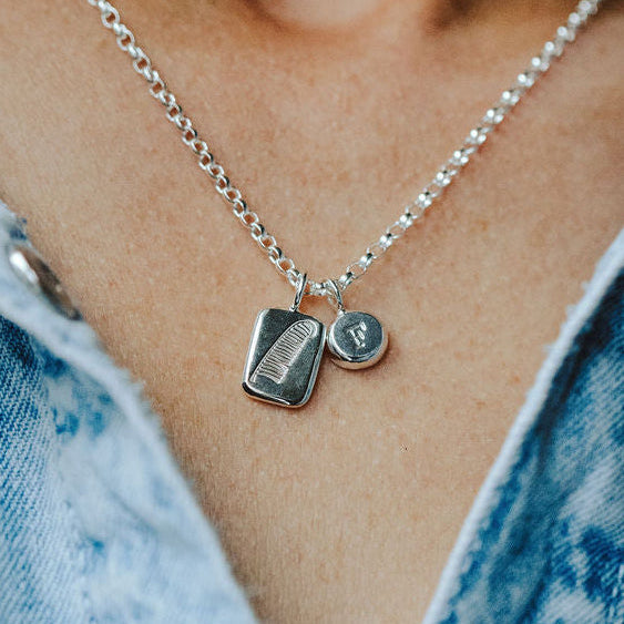 Woman in a denim shirt wearing a necklace with two charm pendants. One pendant has a feather design and the other has an initial on it.