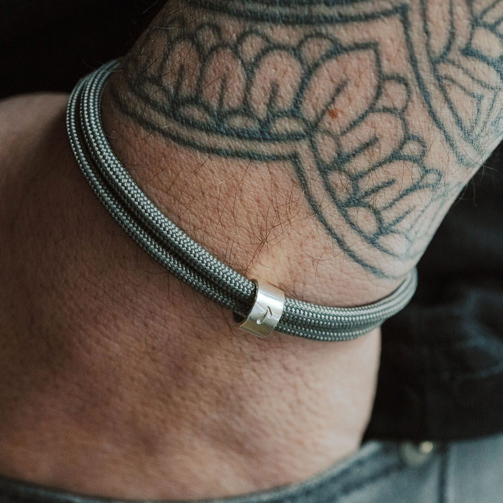 Close-up of a wrist wearing a grey braided bracelet with a silver clasp, against a tattooed arm.