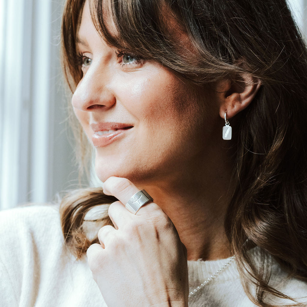 Woman with dark hair and a white knitted t-shirt wearing silver feather charm hoop earrings and a ring stamped with a phoenix design.
