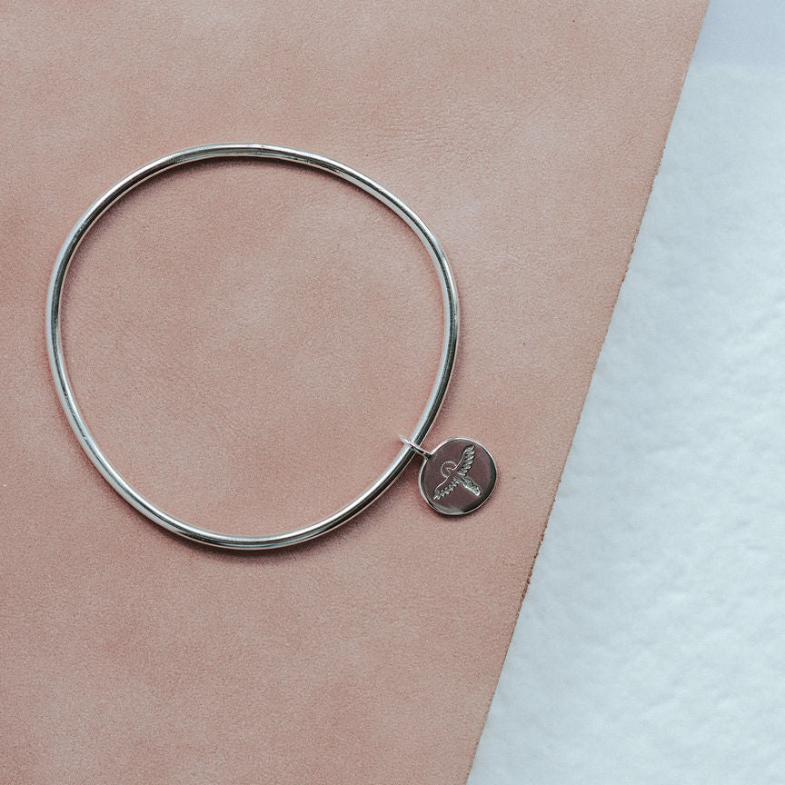 Silver bangle with a small round phoenix charm on a pink surface