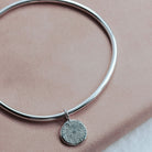 Silver bangle with a round sunrise pendant on a pink surface